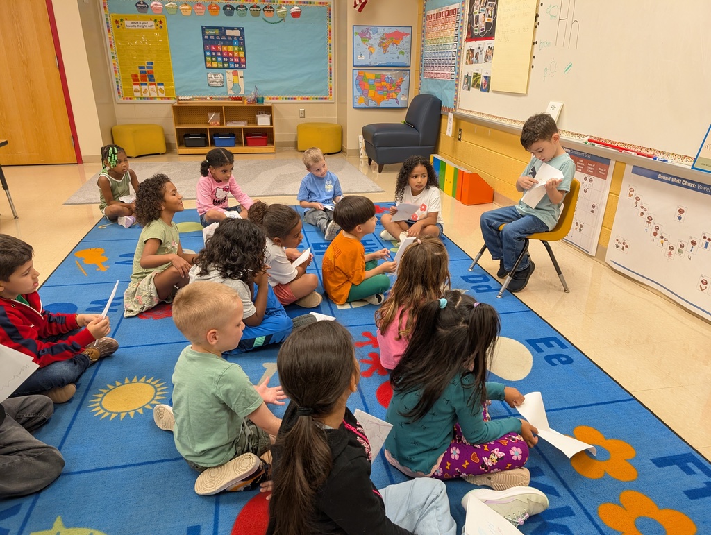 A group of young children sit cross-legged on a colorful classroom rug while one boy sits on a yellow chair at the front, showing his paper to the class. The students on the rug hold their own papers and watch him attentively.