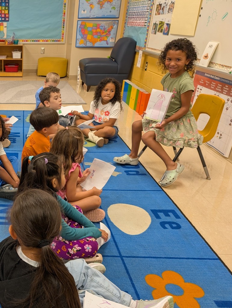 A smiling girl in a green dress and light green shoes sits on a yellow chair at the front of the classroom, holding up her drawing for the class to see. The rest of the children sit on the rug in front of her, holding their own papers and watching.