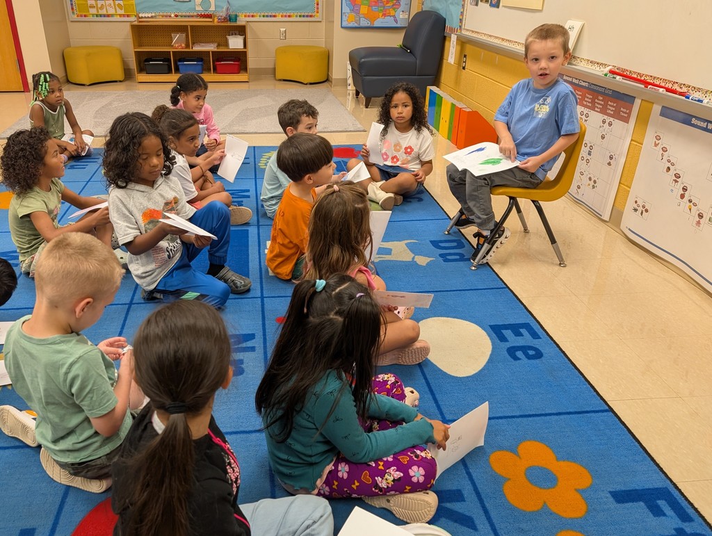 A boy in a light blue shirt sits on a yellow chair at the front of the classroom, holding up his drawing while speaking to his classmates. The children on the rug sit facing him, some looking at his paper and others holding their own drawings.