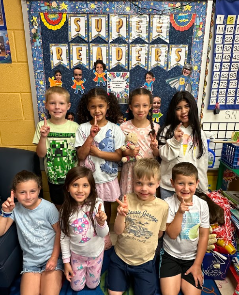 A group of eight smiling children stand in front of the “Super Readers” bulletin board. They hold up one finger to show they are number one, celebrating their reading achievements.