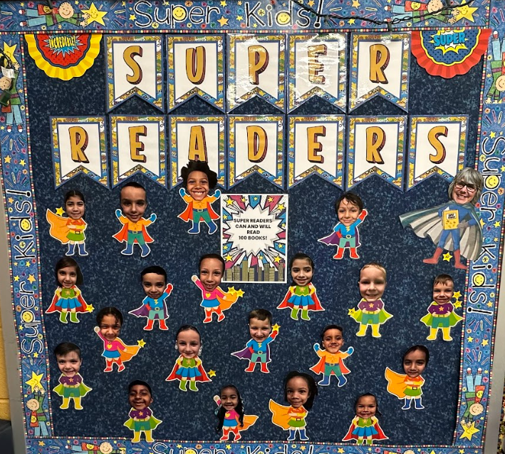 A decorated bulletin board titled “Super Readers” features superhero cutouts with students’ faces added to the characters’ bodies. The display includes the message “Super Readers can and will read 100 books!” surrounded by colorful comic-style decorations.