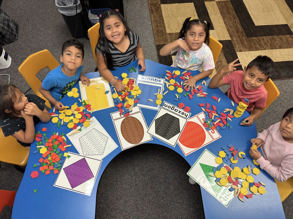 ix young children are seated around a blue, horseshoe-shaped table in a classroom. They are smiling and working with colorful plastic pattern blocks (yellow hexagons, red trapezoids, green triangles, and blue rhombuses). On the table are large laminated shape mats labeled with names like “square,” “hexagon,” “rectangle,” “triangle,” “circle,” and “pentagon.” The children are arranging and stacking the blocks on and around the mats.