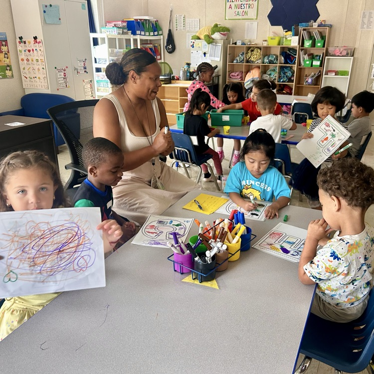 Children sit at tables in a classroom coloring worksheets with markers and crayons. A teacher sits with one group, engaging with the students. One child in the front holds up a scribbled drawing, while others focus on coloring shapes and pictures. The classroom is lively, with cubbies, posters, and supplies visible in the background.