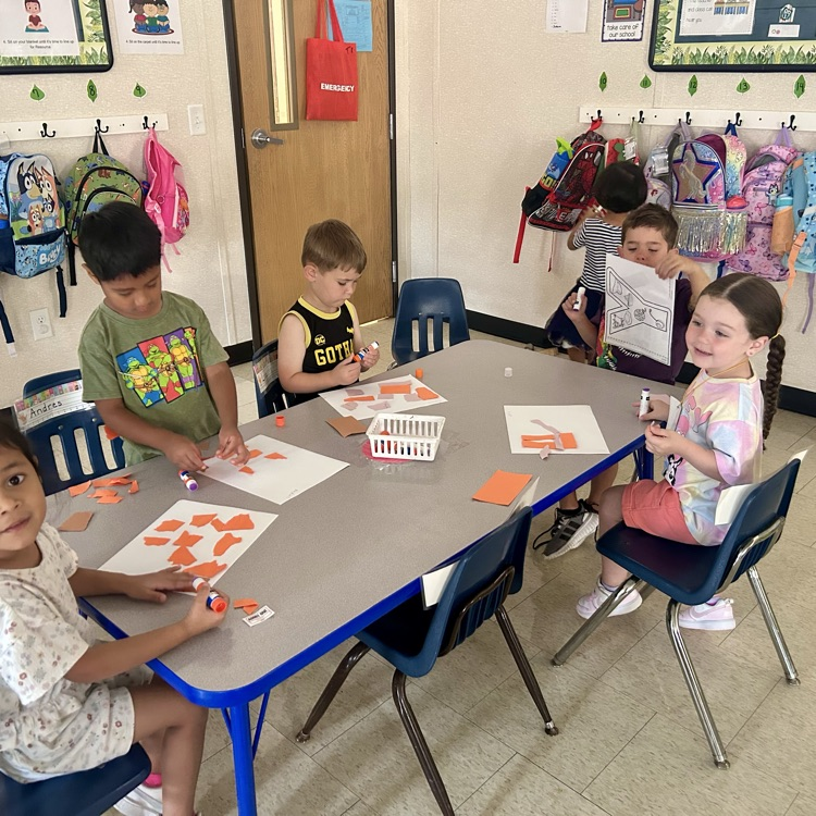 A group of young children sit around a small classroom table working on an art activity. They are gluing orange paper shapes onto white sheets. Backpacks hang on hooks along the wall behind them, and two children stand nearby looking at papers. The classroom is bright and decorated with posters and supplies.