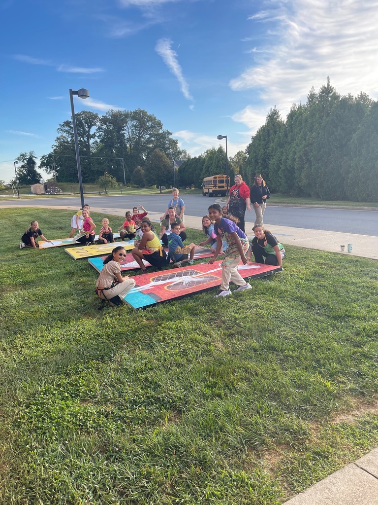 A group of students and adults sit and kneel on the grass outside, painting large wooden flats with bright colors. They are working together on stage scenery under a blue sky, with a school bus and trees in the background.