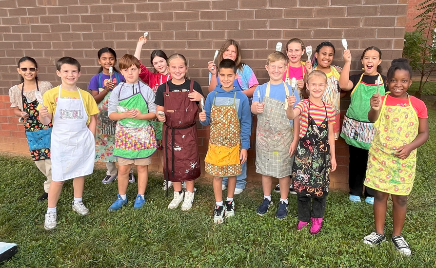 A group of about fifteen children stand outside in front of a brick wall, smiling and holding up paintbrushes. They are dressed in colorful aprons, some with paint smudges, ready to work on stage set pieces for the school musical.