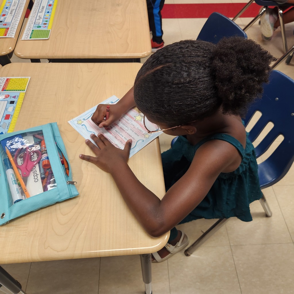 A student with glasses and a green dress sits at a desk, using a red crayon to complete a worksheet. A pencil pouch with markers, scissors, and glue is on the desk.