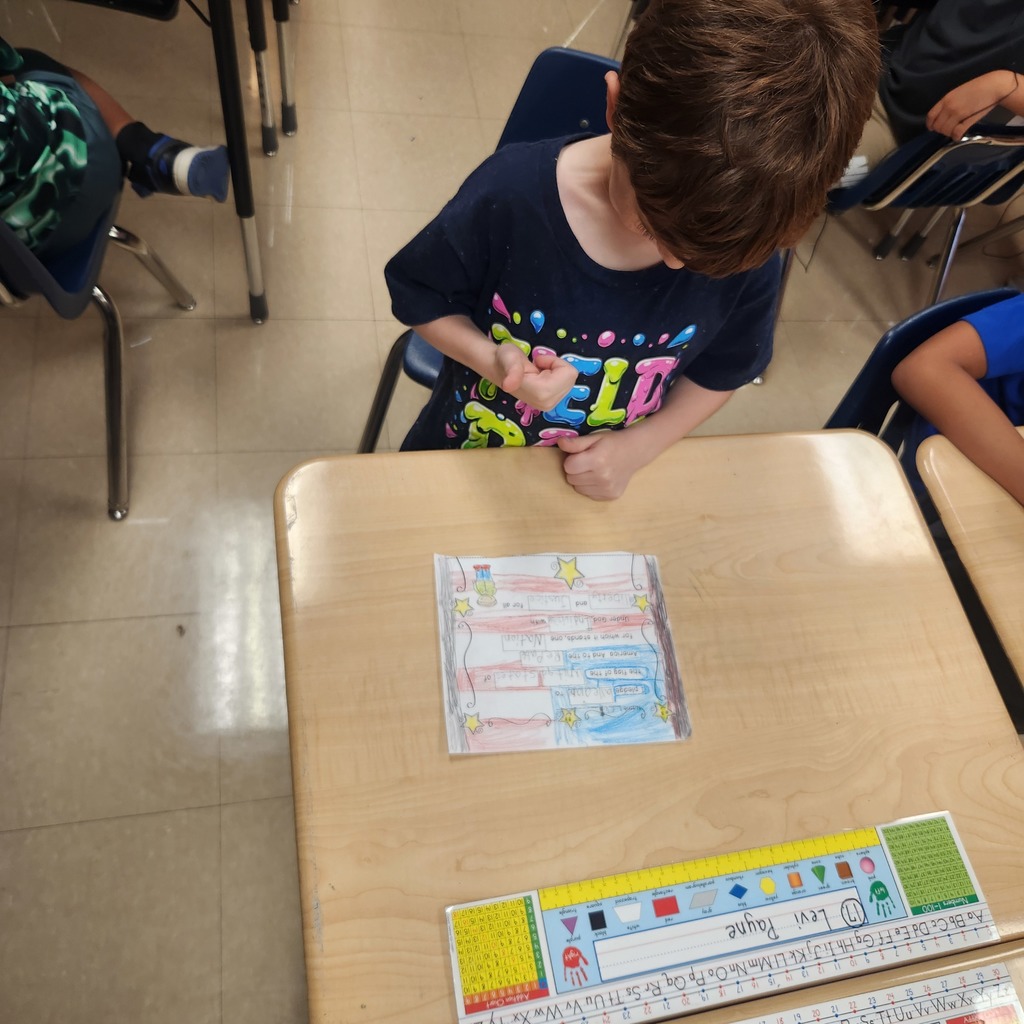 A student in a dark shirt sits at a desk, looking down at a completed worksheet decorated in red, white, and blue with stars. A nameplate and desk ruler are visible in front.