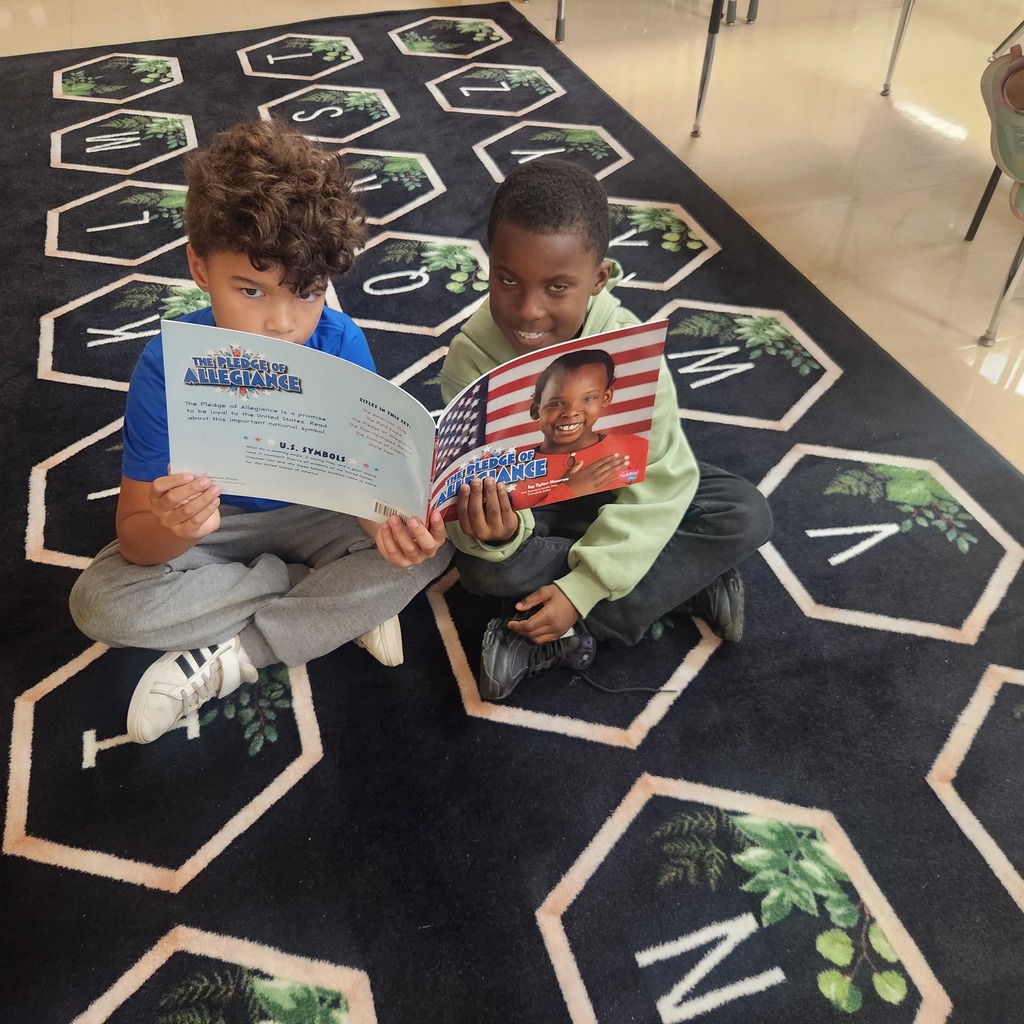 Two students sit cross-legged on a classroom rug with hexagon patterns. They are reading side by side from books titled The Pledge of Allegiance, one holding the front cover showing a smiling child with an American flag background.
