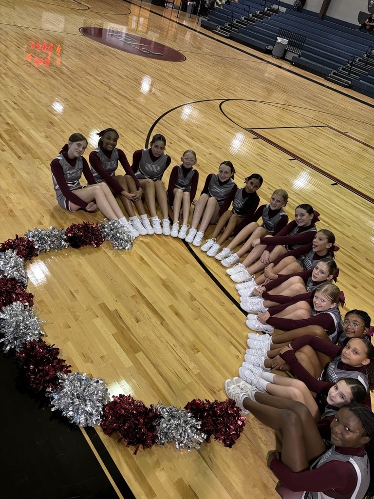 Cheerleaders sit in a circle on a gym floor with their legs extended, creating a heart shape outlined with red and silver pom-poms.