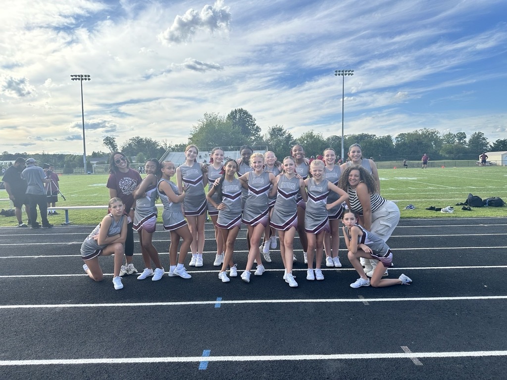 A group of cheerleaders in matching uniforms pose together on a track in front of a football field. They are smiling and making fun poses with their coaches on a sunny afternoon.