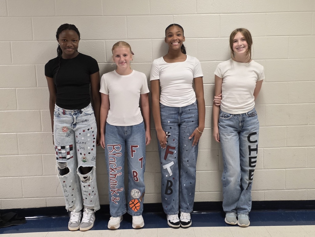 Four students stand in a row against a beige brick wall. From left to right:  The first student wears a black top and ripped jeans decorated with checkerboard patterns, flowers, polka dots, and the words “Game Day” and “Hawks.”  The second student wears a white top and jeans painted with the words “Blackhawks” and “FTB,” along with a football and basketball.  The third student wears a white top and jeans decorated with “FTB” and small dots.  The fourth student wears a white top and jeans with the word “CHEER” painted vertically down one leg.
