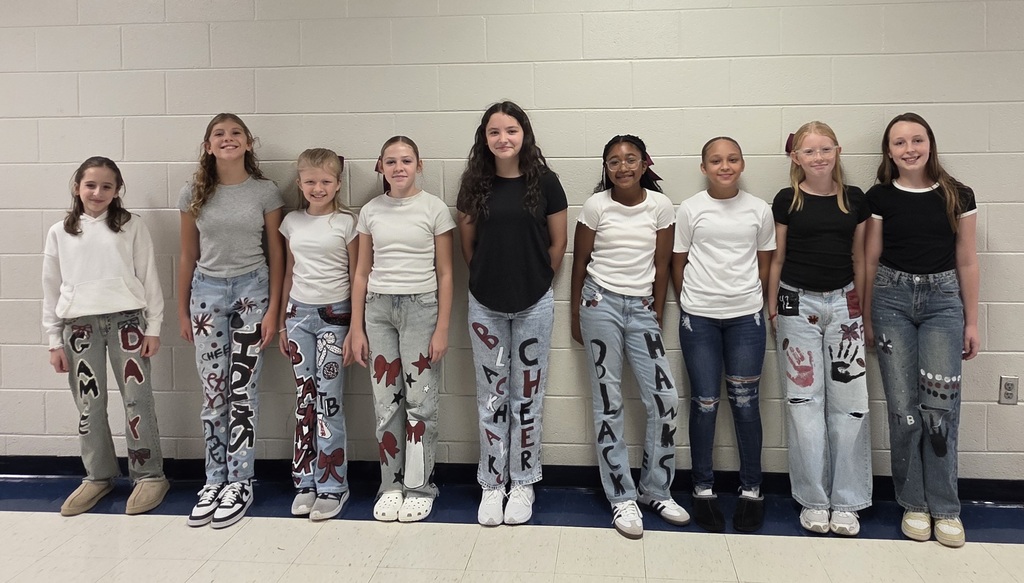 A line of cheerleaders stand indoors against a white wall wearing jeans decorated with hand-painted cheer and Blackhawk designs, paired with plain black or white tops.