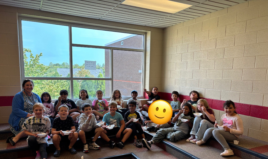 A group of elementary school children sit together on carpeted steps inside a school, smiling and eating bowls of ice cream. A teacher sits on the far left, also smiling. A large window in the background shows trees and part of a brick building outside. The students appear happy and relaxed, enjoying a treat in a casual setting.