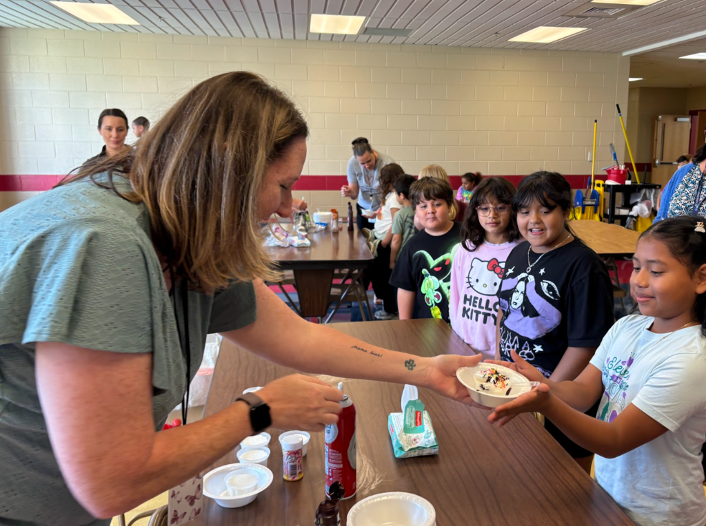 A teacher hands a bowl of ice cream with sprinkles to a smiling student at the front of a line. Several children stand behind her, waiting their turn, while another adult works at a table in the background.