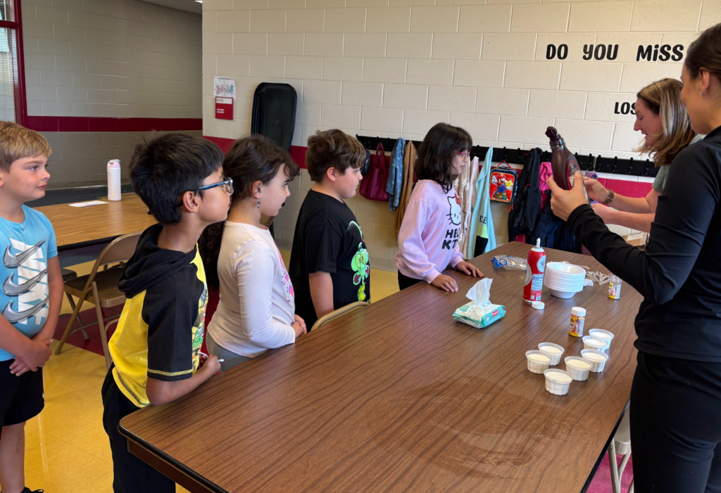 A group of children line up at a table where a teacher is holding a bottle of chocolate syrup, ready to add toppings. The kids watch eagerly, and small cups of toppings and whipped cream are spread out on the table.