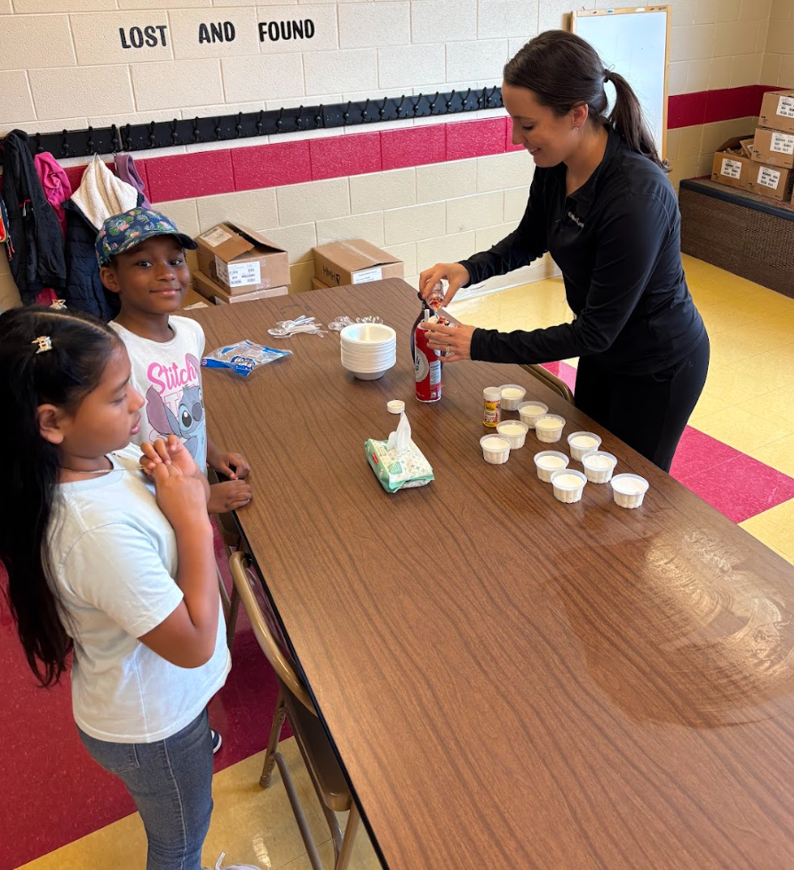 Two students stand at a table where a teacher is adding whipped cream to bowls of ice cream. Small cups of toppings and supplies are lined up across the table, and the children smile as they wait.