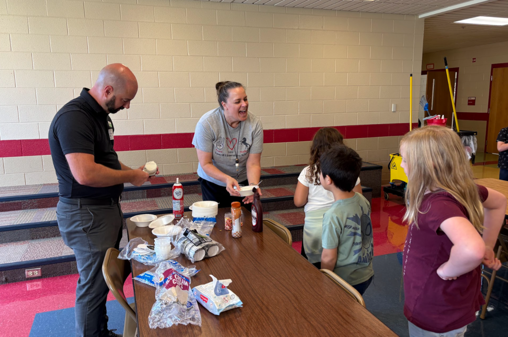 Two teachers serve ice cream and toppings to a group of children waiting at a table. One teacher laughs while holding a bowl of ice cream, and the other prepares a serving. Containers of supplies and toppings are scattered on the table.