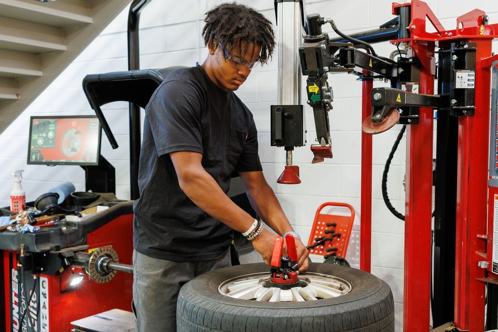 A student in safety glasses operates a red tire changing machine, adjusting a wheel as part of automotive training.