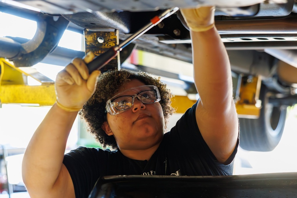 A student in safety glasses uses a ratchet tool while working underneath a raised vehicle in an automotive shop.
