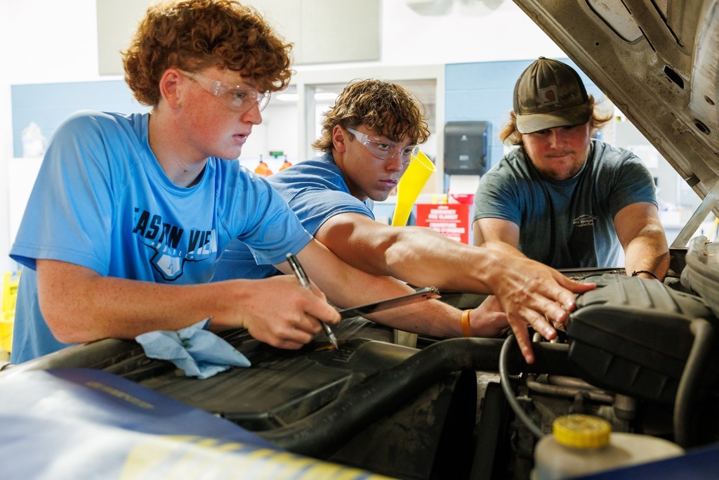 Three students in safety glasses lean over the open hood of a car, using tools and working together on the engine during class.