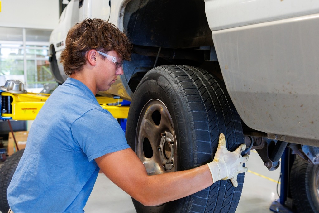 A student wearing safety glasses and gloves lifts and positions a tire onto the wheel hub of a vehicle raised on a yellow hydraulic lift.