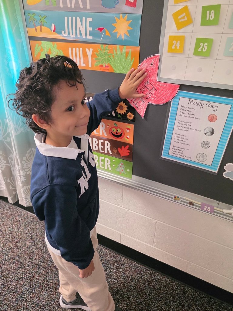 A smiling boy wearing a navy blue sweater places a red fish cutout on the classroom calendar board