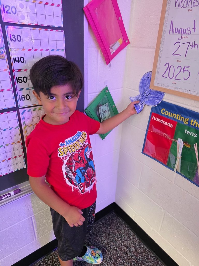 A boy in a red Spider-Man shirt holds a blue fish cutout next to a classroom number chart and calendar.