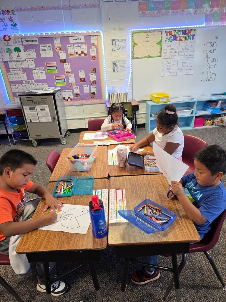 Four students at a table work on coloring and cutting fish cutouts, with supply boxes and crayons scattered on the desks.