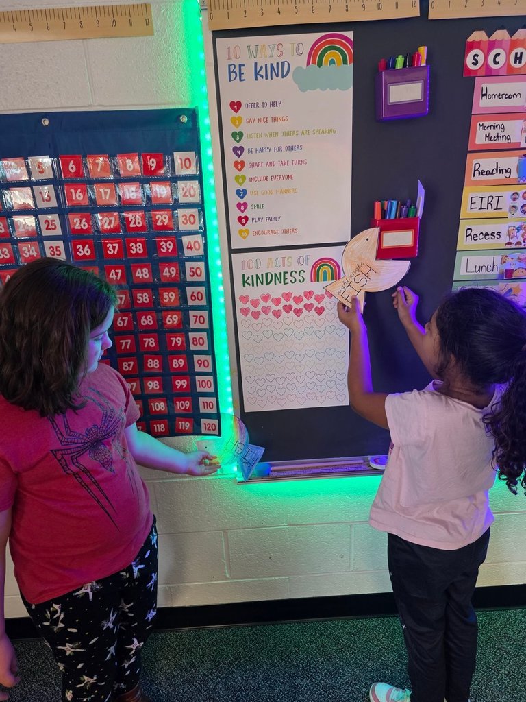 Two girls stand by a classroom kindness poster; one is taping a paper fish to the wall.