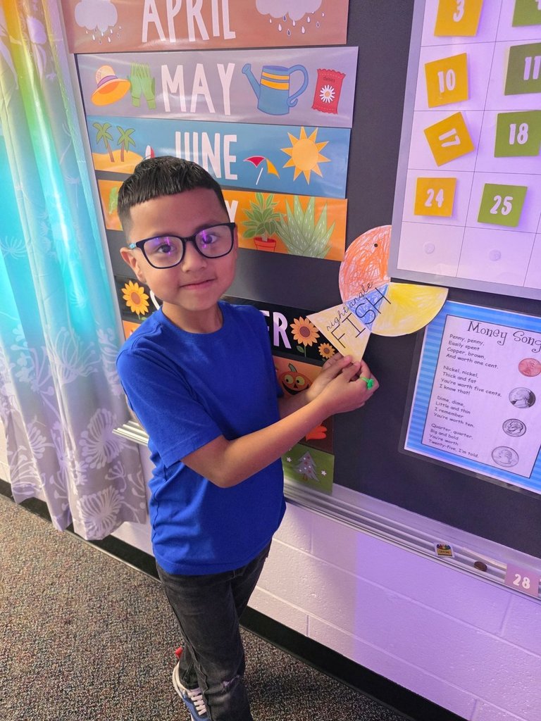 A boy in a blue shirt and glasses places an orange and yellow fish cutout on the classroom board.