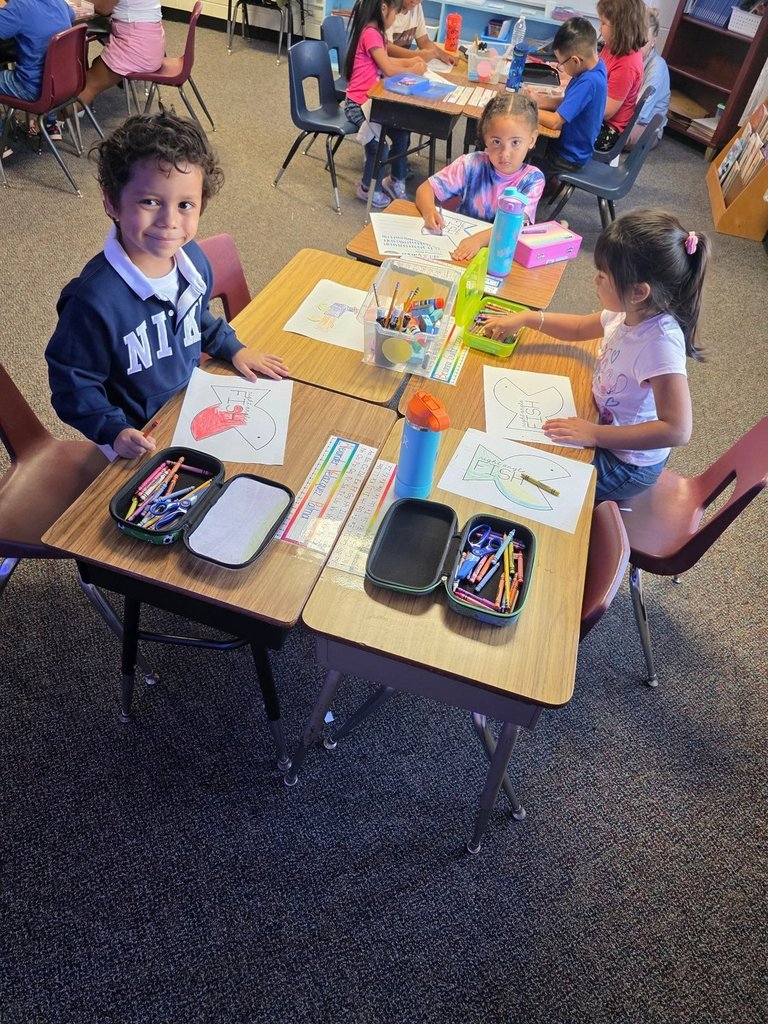 A group of four students at a table coloring paper fish cutouts, with one smiling at the camera holding a red fish.