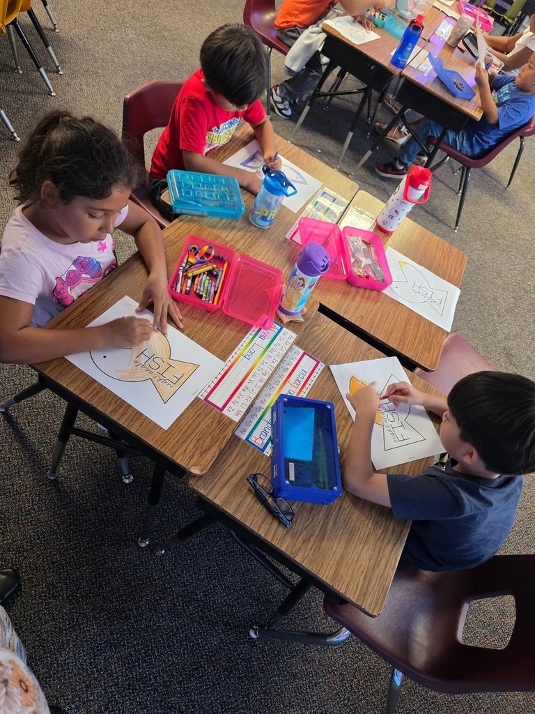 Three students sit at desks coloring paper fish cutouts with crayons, with pencil boxes and water bottles on their desks.