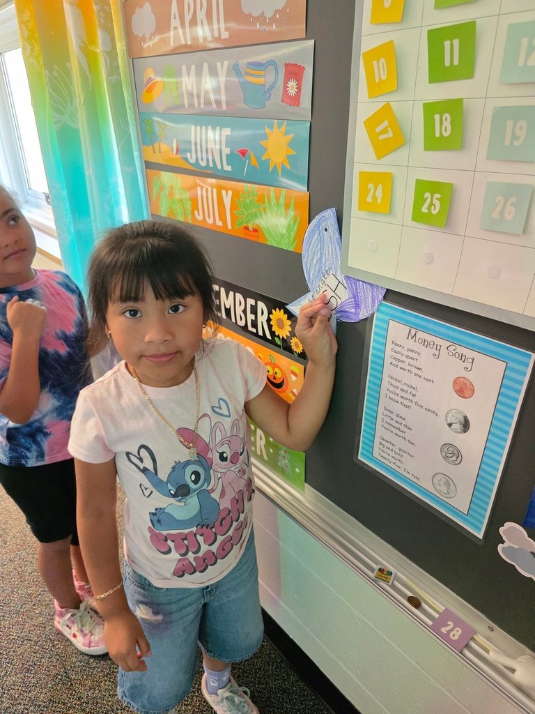 A young girl wearing a Stitch and Angel shirt holds up a blue paper fish labeled right angle fish next to a classroom calendar display.