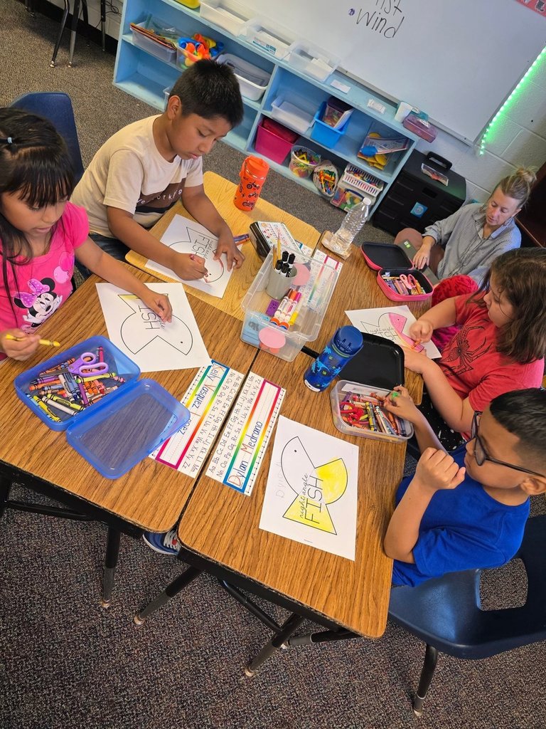 Five students sit at a table coloring fish cutouts, with a teacher sitting nearby assisting.