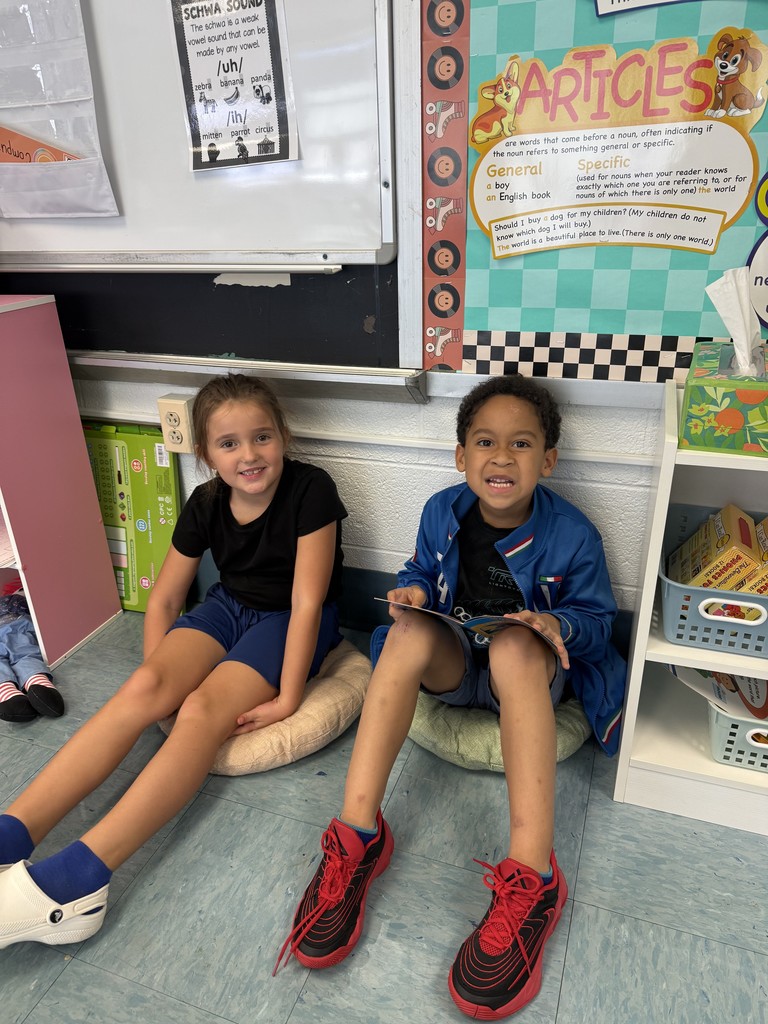 Two children sit on cushions against the classroom wall near a poster about articles, one holding a book while both smile.