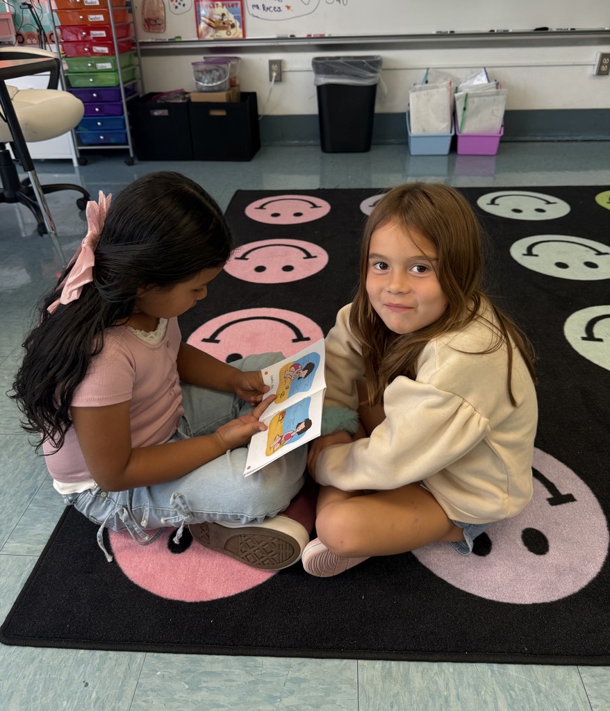 Two girls sit on a rug with smiley faces, one holding an open book while the other looks toward the camera.
