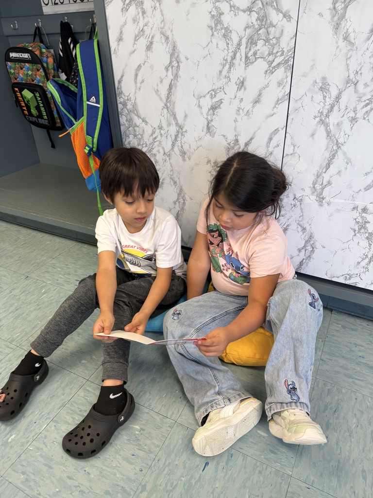 Two children sit on the floor near cubbies, reading a book together.