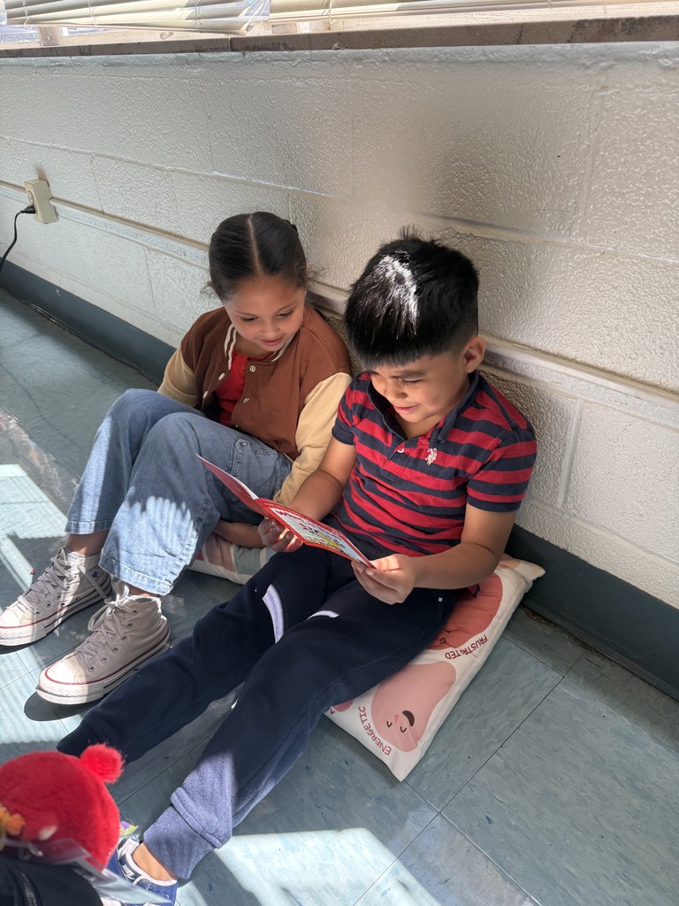 A girl and a boy sit against a classroom wall, the boy reading a book aloud while the girl listens.