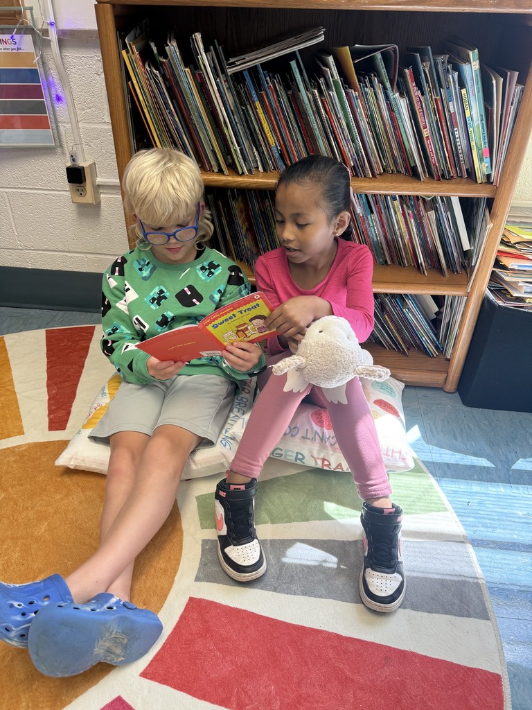 A girl and a boy sit together on cushions in front of a bookshelf, reading a book together; the girl holds a stuffed toy.