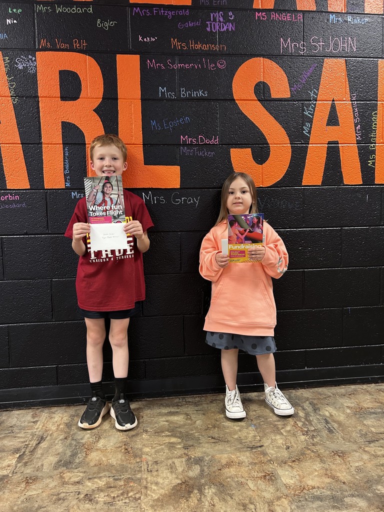 Two students hold their Urban Air passes while standing in front of a black wall with orange lettering and teacher names written in colorful chalk.