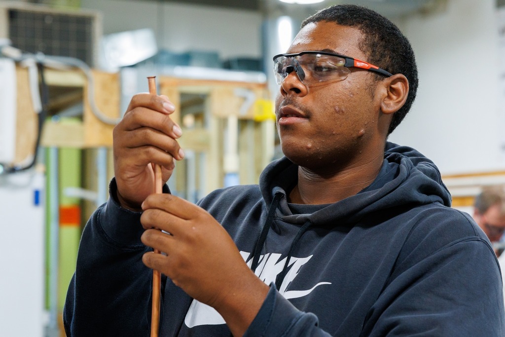A student in safety glasses holds up a piece of copper tubing, inspecting the flared end during HVAC training.