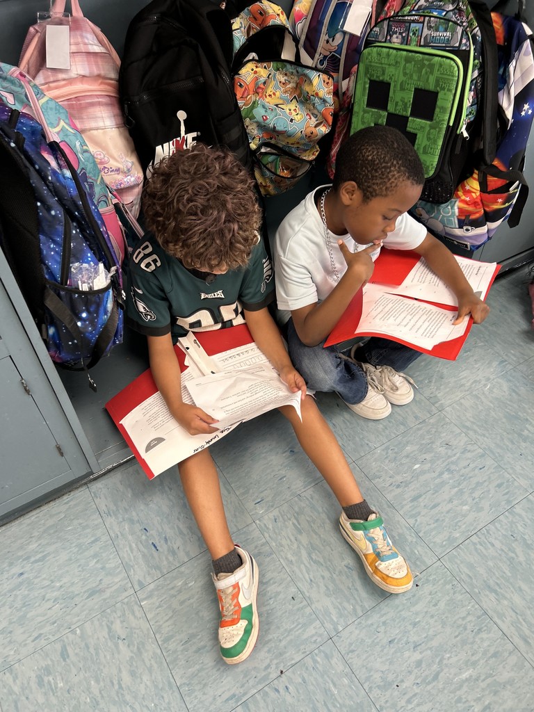 Two children sitting by classroom lockers with backpacks behind them, reading from red folders.