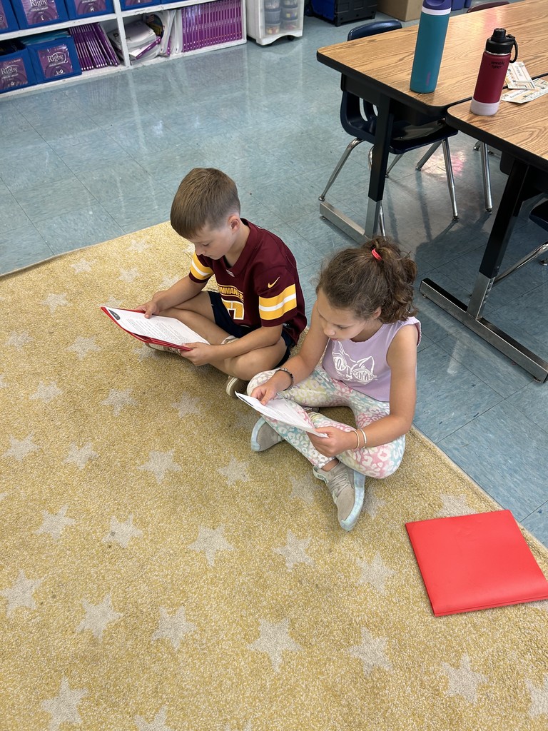 Two children sitting on a rug, reading from papers inside red folders.