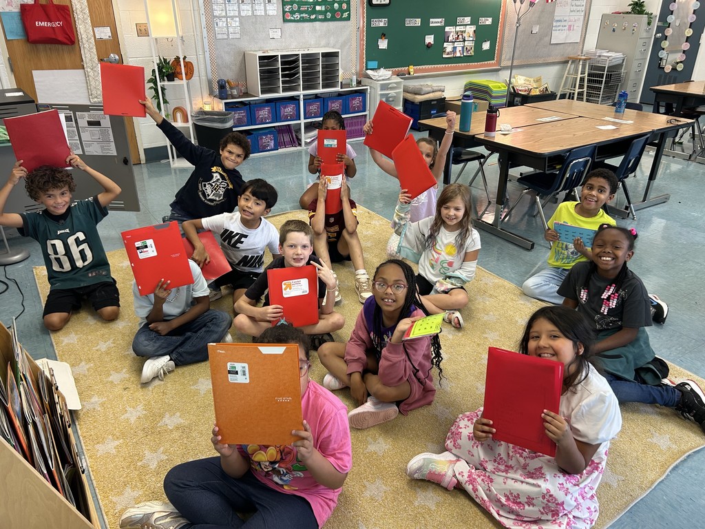 A group of elementary students sitting on a classroom rug, smiling and holding up red folders.