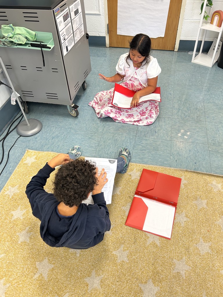 Two children sitting on the floor, facing each other, reading from red folders.