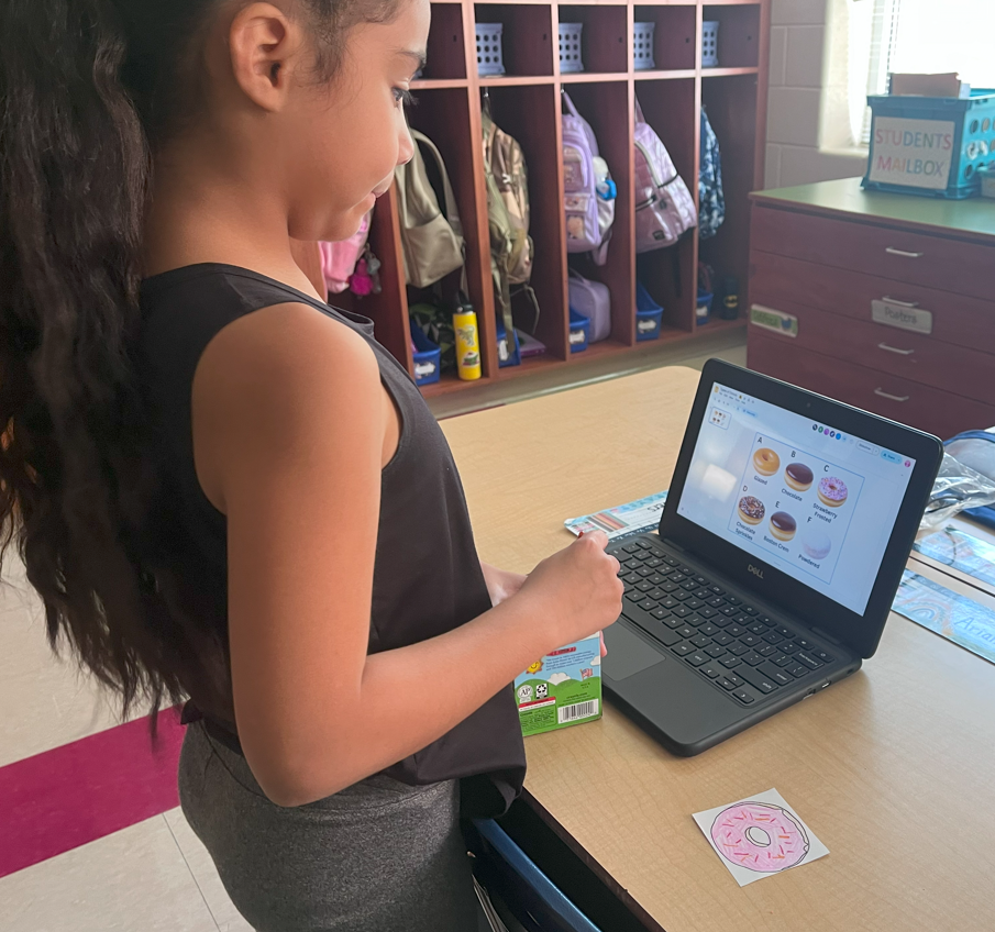 A student stands at a desk looking at a laptop screen showing images of donuts with labels. A paper donut cutout is on the desk in front of her. Classroom cubbies with backpacks are in the background.