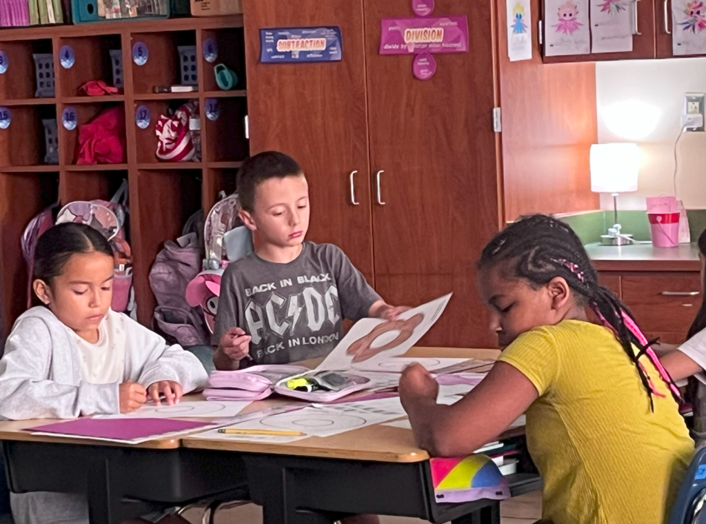 Three students at a shared table work on coloring and cutting out paper donuts. A boy in an AC/DC shirt holds up his donut while others continue coloring. Classroom cubbies and backpacks are visible in the background.