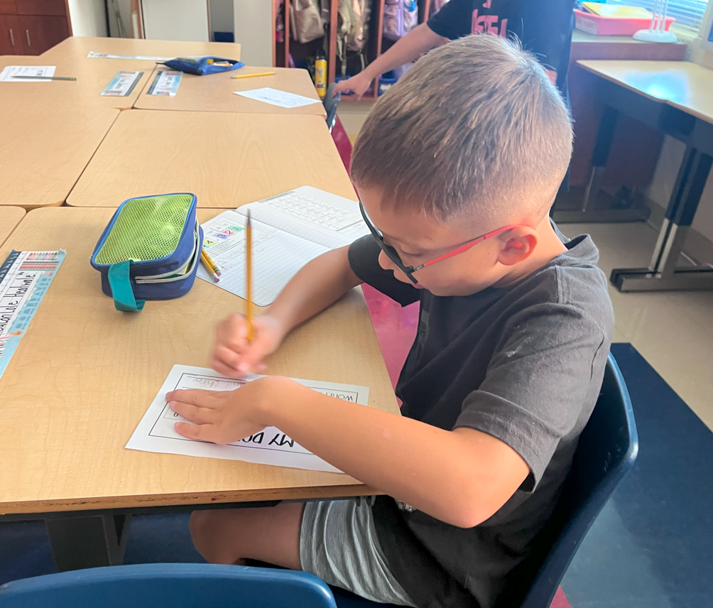 A student wearing glasses sits at a desk writing on a worksheet labeled “My Donut.” He is focused, holding a pencil, with a notebook and pencil case nearby.