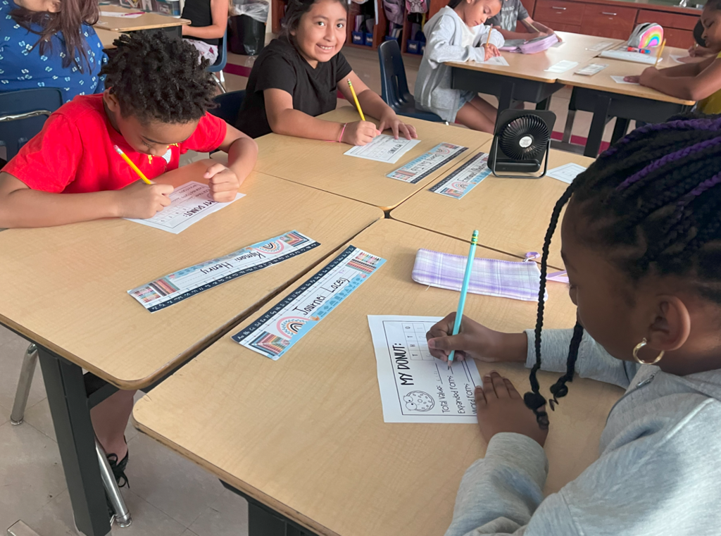 Several students sit at desks working on the “My Donut” math activity, writing with pencils. A girl in the foreground is filling in her paper, while another student smiles at the camera.